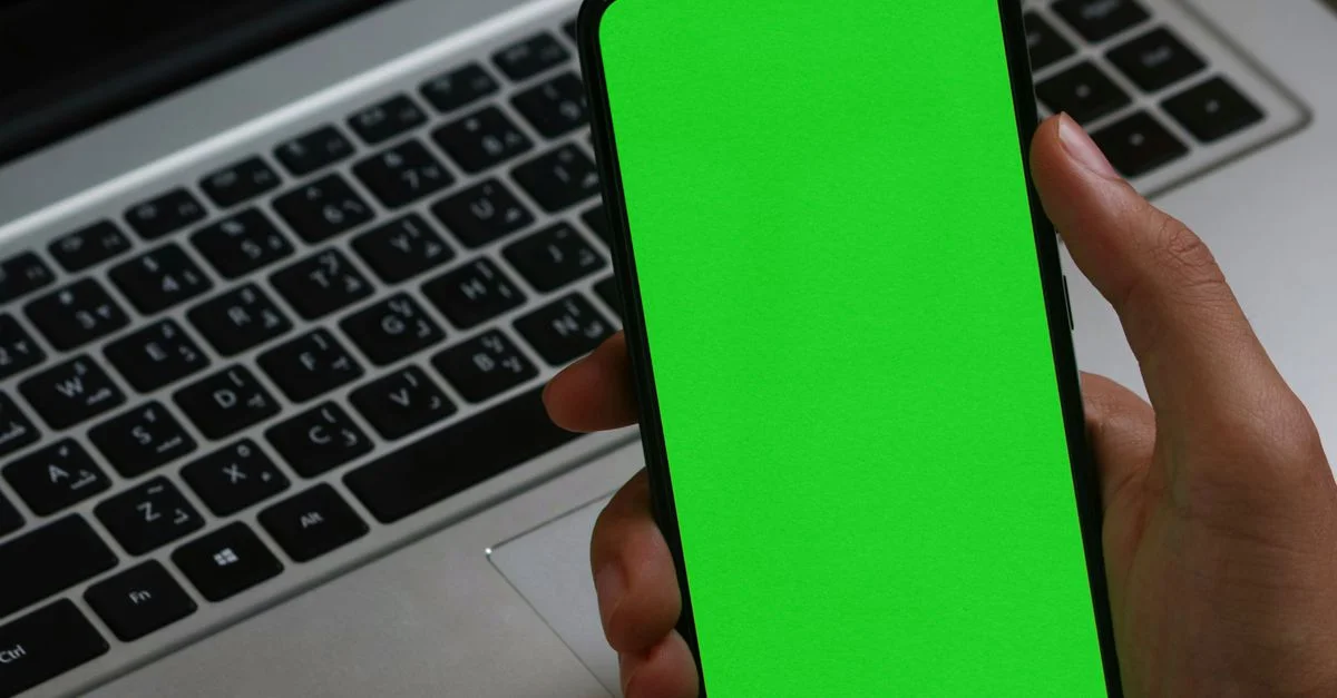 Une main tenant un smartphone avec un écran vert superposé à un ordinateur portable, idéal pour les présentations de maquettes numériques.