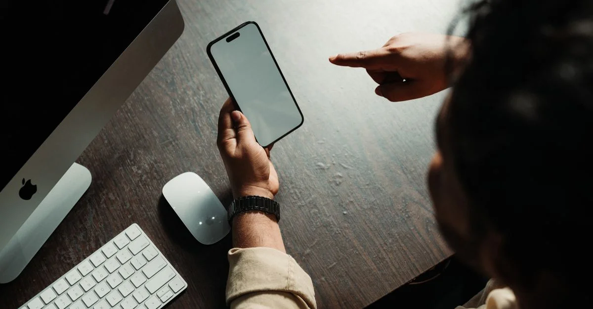 Personne utilisant un smartphone à un bureau avec un ordinateur et des périphériques.