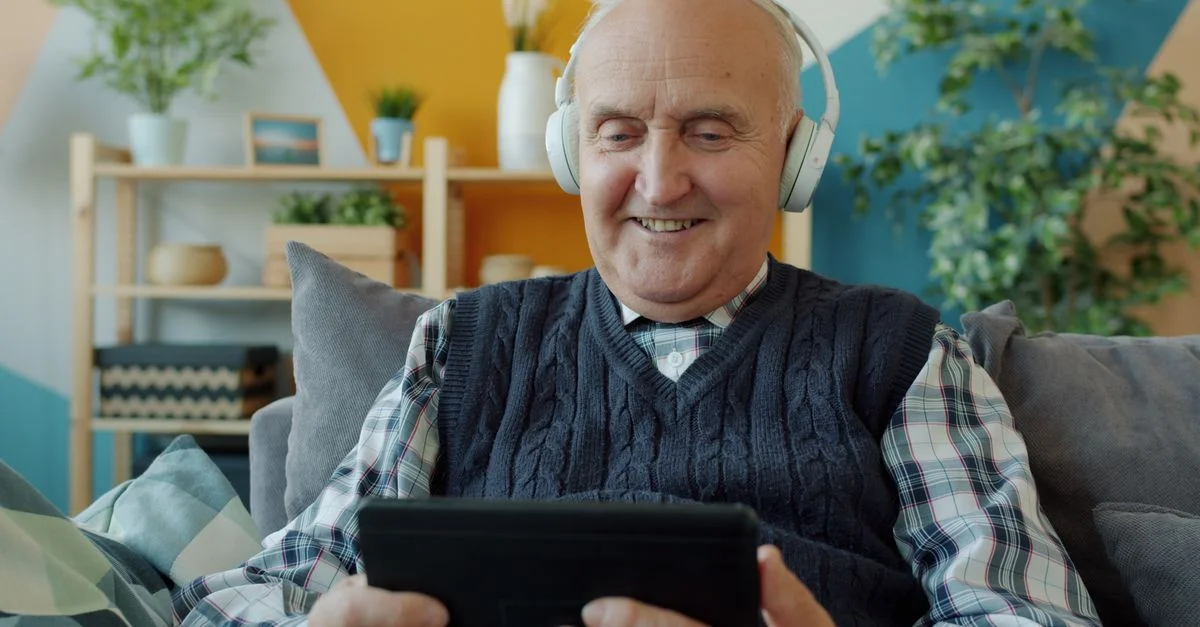Un homme âgé utilise des écouteurs et une tablette, se détendant sur un canapé à l'intérieur.