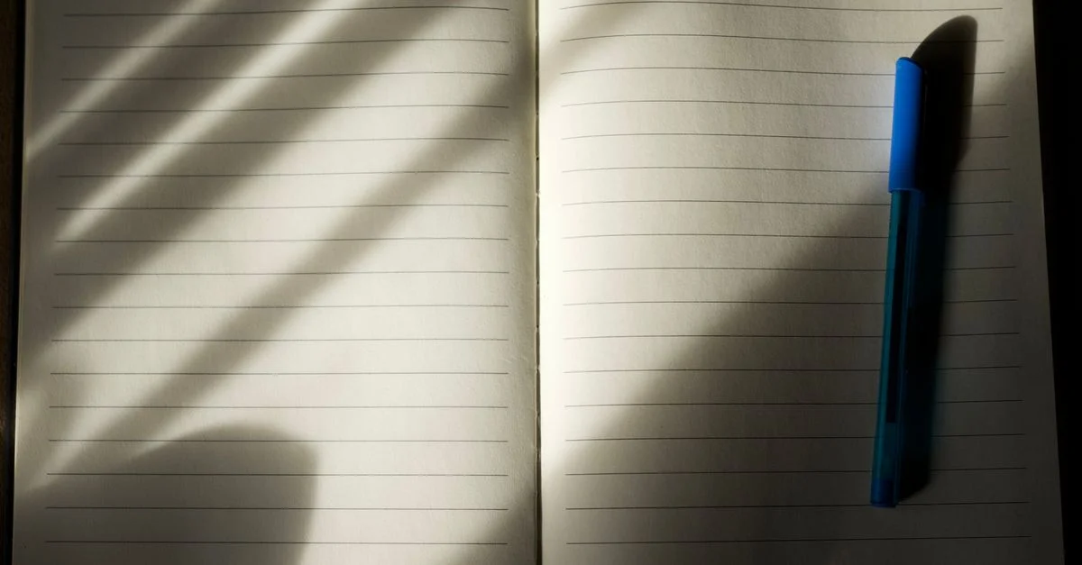 Un carnet à lignes ouvert avec un stylo bleu sous une douce lumière naturelle projetant des ombres légères.