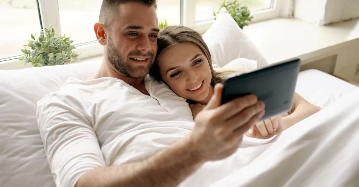 Couple heureux se détendant au lit en regardant une tablette ensemble à l'intérieur.