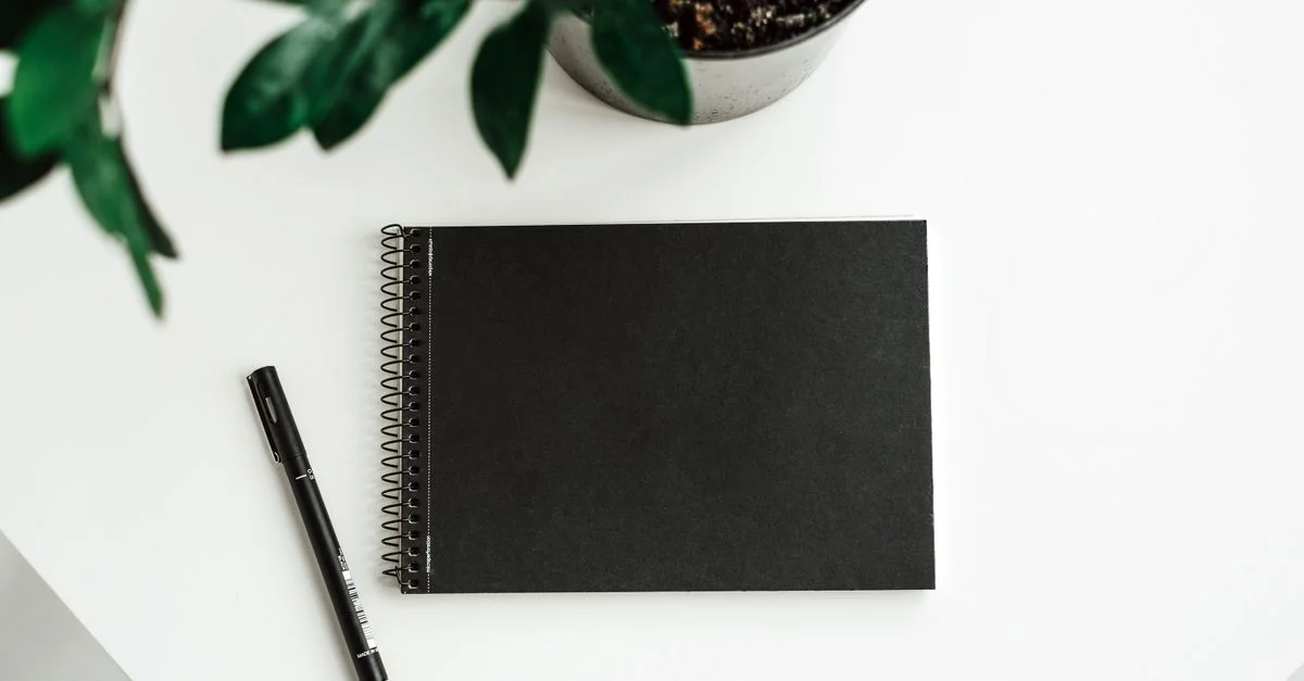 Mise en page minimaliste mettant en vedette un carnet noir, un stylo et une plante sur une table blanche, parfait pour les créatifs.