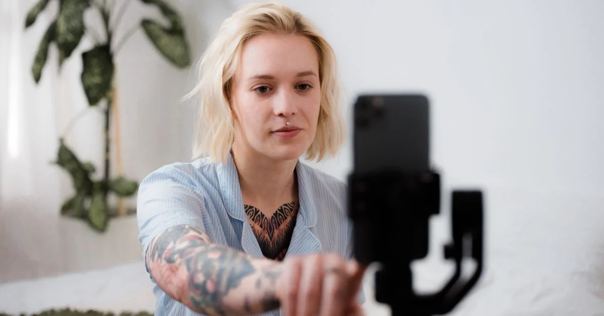 Femme tatouée aux cheveux blonds ajuste un smartphone sur un trépied pour préparer un vlog à l'intérieur.